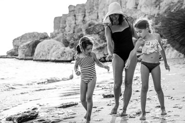 Anne ve iki küçük kız, deniz kıyısında mayo giyip kuma bakarak yürüyorlar. Aile deniz tatili.