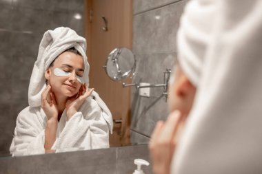 Kafasında havluyla bornoz olan bir kız banyoda aynanın önünde gözlerinin altına yamalar yapıştırıyor..