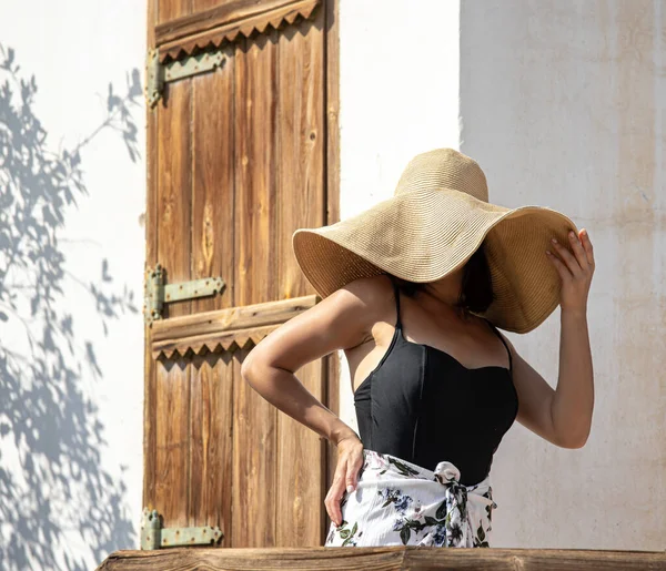 A girl in a big straw hat on the porch of an old house on a sunny hot day.