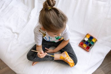 Çocuk bir fırçayla ayağına bir desen çiziyor. Üzerinde parlak renkler olan komik bir çizim..