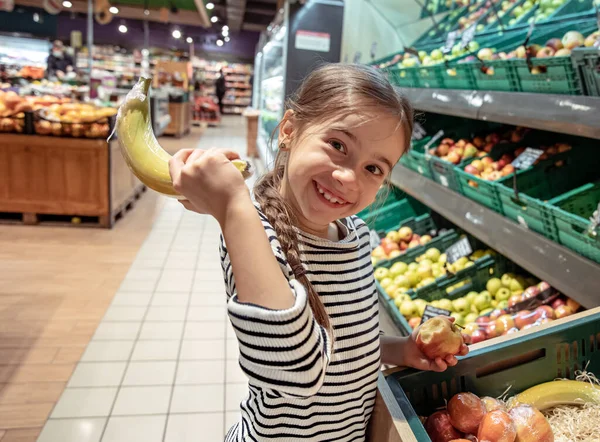 Funny little girl with banana at the supermarket. Individual packaging of fruits.