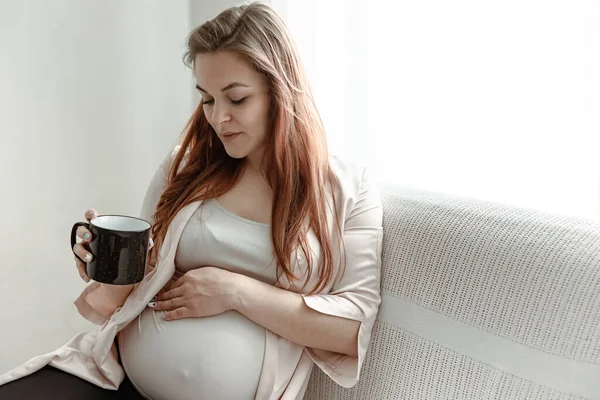 大肚子的时髦孕妇坐在家里的沙发上喝茶或喝咖啡 — 图库照片