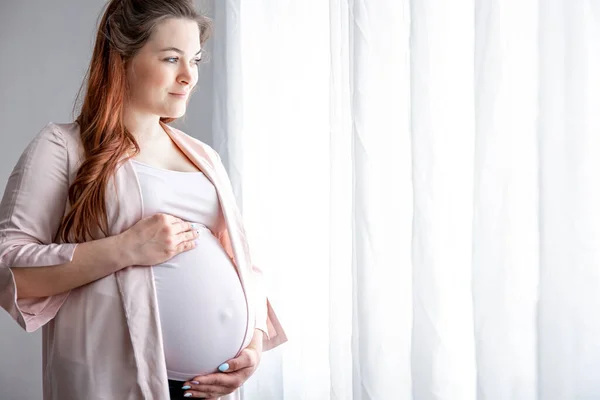 Stylish pregnant woman in the last months of pregnancy stands near the window.