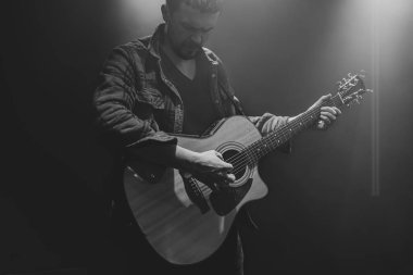 Bir adam kısmen aydınlatılmış bir konserde akustik gitar çalıyor..