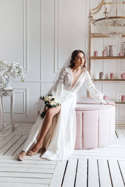 Parlak bir odada bir buketle poz veren güzel bir boho gelinlik içinde çekici genç bir gelin. Düğün fotoğrafı, gelin sabahı..