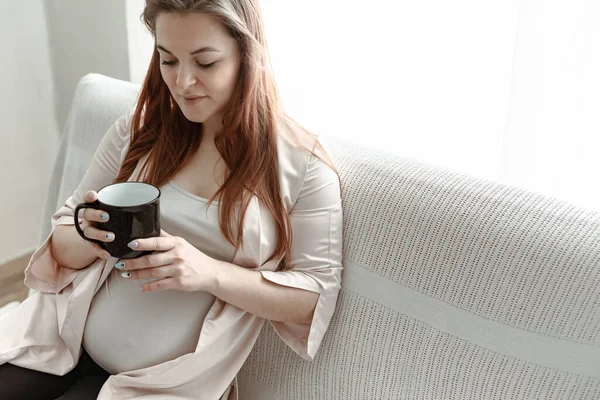 大肚子的时髦孕妇坐在家里的沙发上喝茶或喝咖啡 — 图库照片
