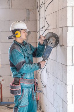 Bir iş tesisinde koruyucu miğfer takmış elektrikçi inşaat işçisi bir öğütücüyle çalışıyor..