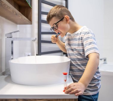 Küçük şirin bir çocuk dişlerini fırçalıyor ve saati kum saatiyle tutuyor..