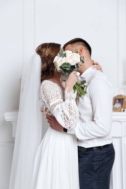 Gelin ve damat bir buket çiçeğin arkasına saklanıp öpüşüyorlardı. Sıcak bir odada düğün fotoğrafı çekimi..