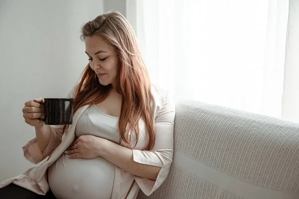 Stylish pregnant woman with a big belly sits on the sofa at home and drinks tea or coffee.