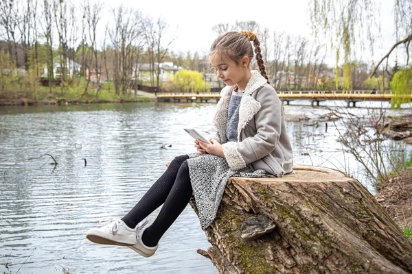 A little girl checks her phone, sitting by the river, and does not pay attention to the nature around.