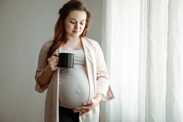 一个大肚子 手里拿着一杯茶或咖啡的时髦孕妇站在窗边 — 图库照片