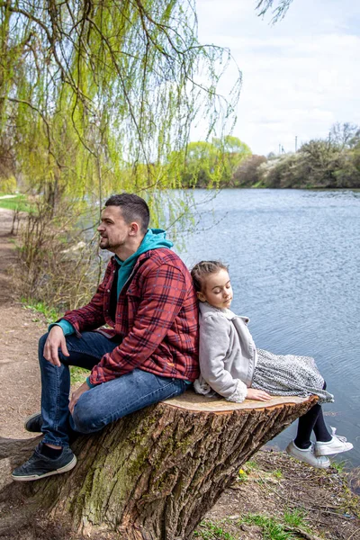 Dad and his little daughter sit in the forest by the river in early spring and enjoy nature.