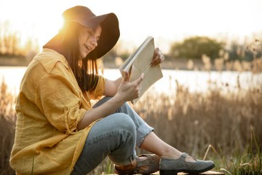Yüzünde gülümseme olan genç bir kadın gün batımında nehrin kenarında oturmuş kitap okuyor..
