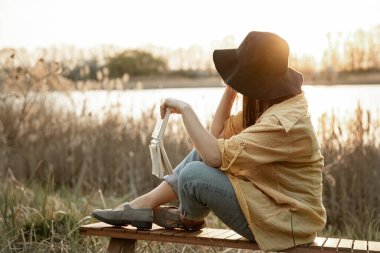 Şapkalı şık kız, gün batımında nehrin kenarında otururken arkadan kitap okuyor..