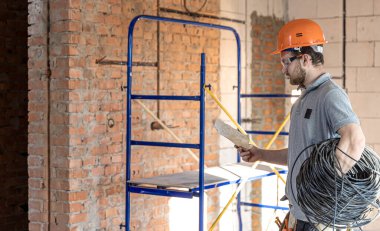 Bir elektrikçi, şantiyede elinde bir elektrik kablosu tutarken bir inşaat çizimini inceliyor..