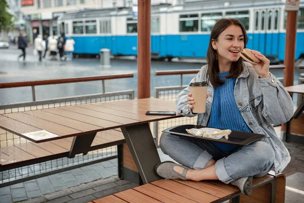 평상복 차림의 매력적 카페의 여름철 테라스에 커피를 마시며 햄버거를 먹는다 — 스톡 사진