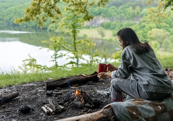 Young woman resting near the fire with a cup of warming drink in the forest near the river.