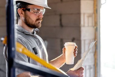 Bir elektrikçi elinde kahveyle bir inşaat çizimini inceliyor..