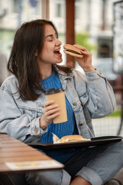 Gündelik tarzda çekici bir kız yaz terasında oturmuş kahveli burger yiyor..