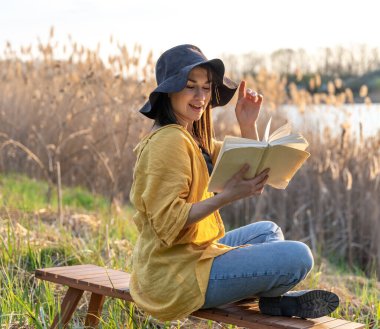 Şapkalı çekici kız gün batımında doğada kitap okuyor..