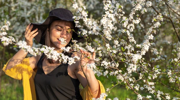 春の花の香りを楽しむ花の木の間の帽子の魅力的な女の子 — ストック写真