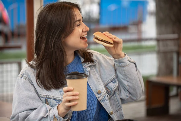 평상복 차림의 매력적 소녀가 여름철 테라스에 커피와 햄버거를 먹는다 — 스톡 사진