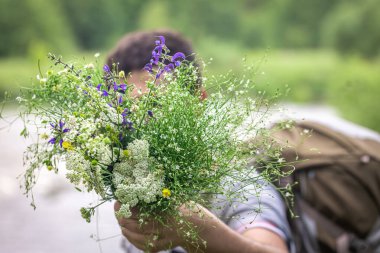 Yürüyüşe çıkmış bir adam arka planda bir buket kır çiçeği tutuyor..