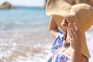 Şapkalı çekici kız yüzünü güneşten saklar, deniz kıyısında oturur..