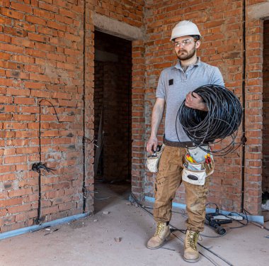 Sert şapkalı bir elektrikçi elinde elektrik kablosu tutarken duvara bakar..
