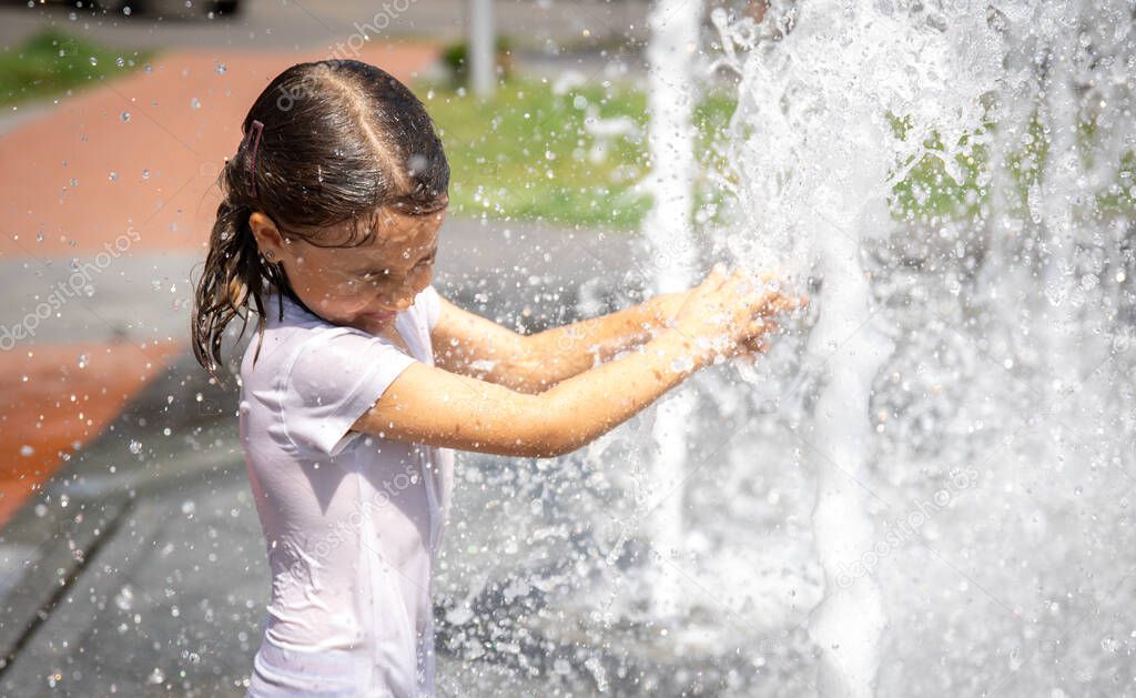 Happy little girl among the splashing water of the city fountain has fun and escapes from the heat.