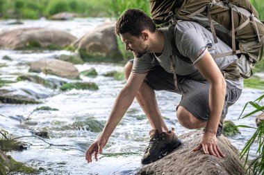 Büyük bir sırt çantası olan bir turist yaz sıcağında bir dağ nehrinin yanında serinliyor..