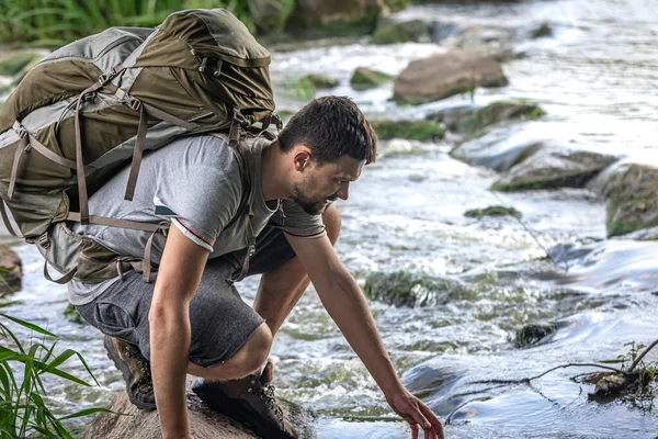 Büyük bir sırt çantası olan bir turist yaz sıcağında bir dağ nehrinin yanında serinliyor..