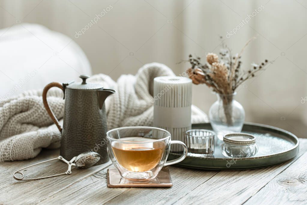 Cozy still life with a glass cup of tea, a teapot and candles on a blurred background.