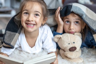 Küçük kız kardeşler odada yerde yatan oyuncak ayıyla kitap okuyorlar..