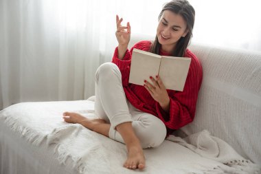 Kırmızı kazaklı çekici genç kadın kanepede otururken kitap okuyor..