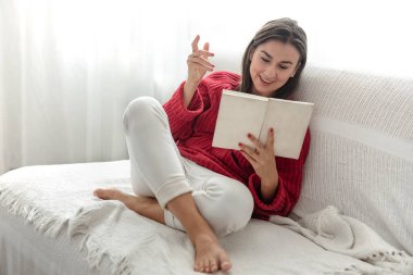 Kırmızı kazaklı çekici genç kadın kanepede otururken kitap okuyor..