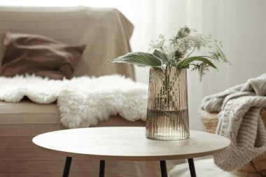 Masanın üzerinde bitkiler olan cam vazo, İskandinav tarzında oturma odasının bulanık arka planına karşı.