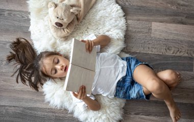Çocuk evde rahat bir halının üzerinde en sevdiği oyuncak ayısıyla uzanan bir kitap okuyor..