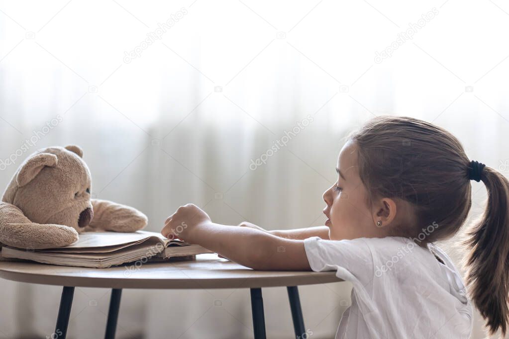 A little girl plays with her teddy bear and a book, teaches him to read, plays at school.