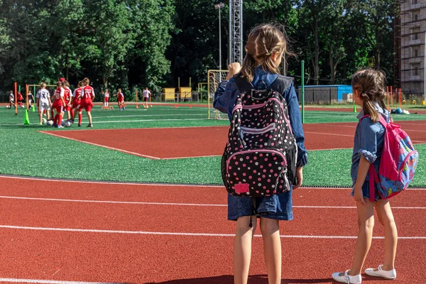小さな女の子の学校の子供たちはスタジアムでバックパックを着て少年たちがサッカーを見て — ストック写真