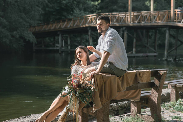 A beautiful young woman with flowers and her husband are sitting on a bench and enjoying communication, a date in nature, romance in marriage.
