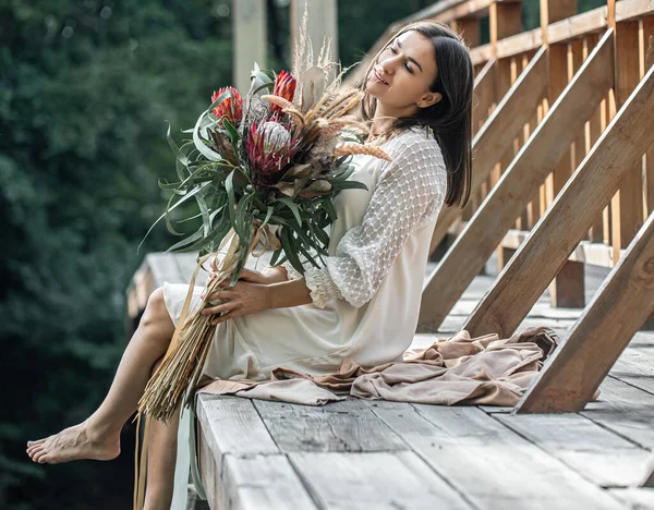 白いドレスの若い女性がエキゾチックな花の花束で木製の橋の上に座っています — ストック写真