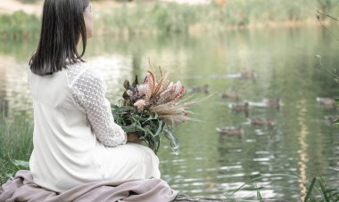 Beyaz elbiseli esmer bir kız nehir kenarında oturur. Bir buket egzotik çiçek, bulanık bir arka plan, arka plan....