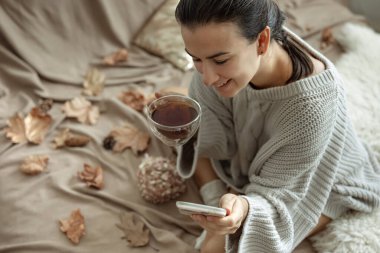 Bir kadın telefonu kullanıyor ve bir fincan çay tutuyor. Yatakta rahat örülmüş bir süveterle oturuyor. Arka planı sonbahar yapraklarıyla bulanık..