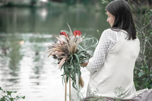 白いドレスのブルネットの女の子はエキゾチックな花の花束で川のそばに座って 背景をぼかし バックビュー — ストック写真