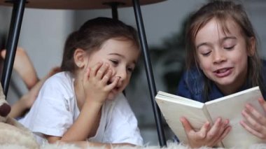 Küçük kız kardeşler evde yerde uzanmış kitap okurken eğleniyorlar..