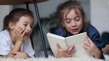 Küçük kız kardeşler evde yerde uzanmış kitap okurken eğleniyorlar..