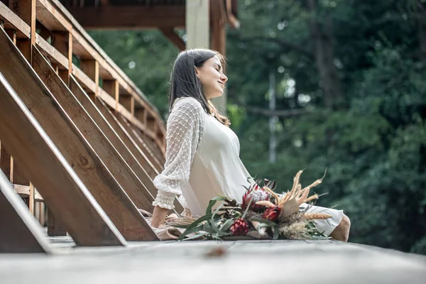 白いドレスの若い女性がエキゾチックな花の花束で木製の橋の上に座っています — ストック写真