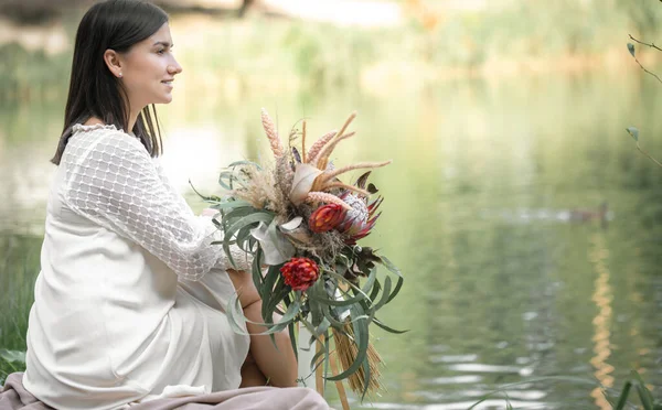 白いドレスのブルネットの女の子はエキゾチックな花の花束で川のそばに座って 背景をぼやけて — ストック写真